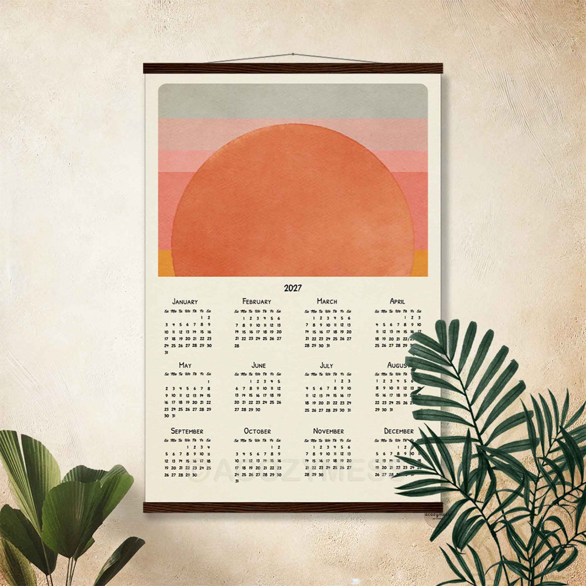 2027 sunset wall calendar in a dark wood hanger frame on a textured beige wall with palm leaves