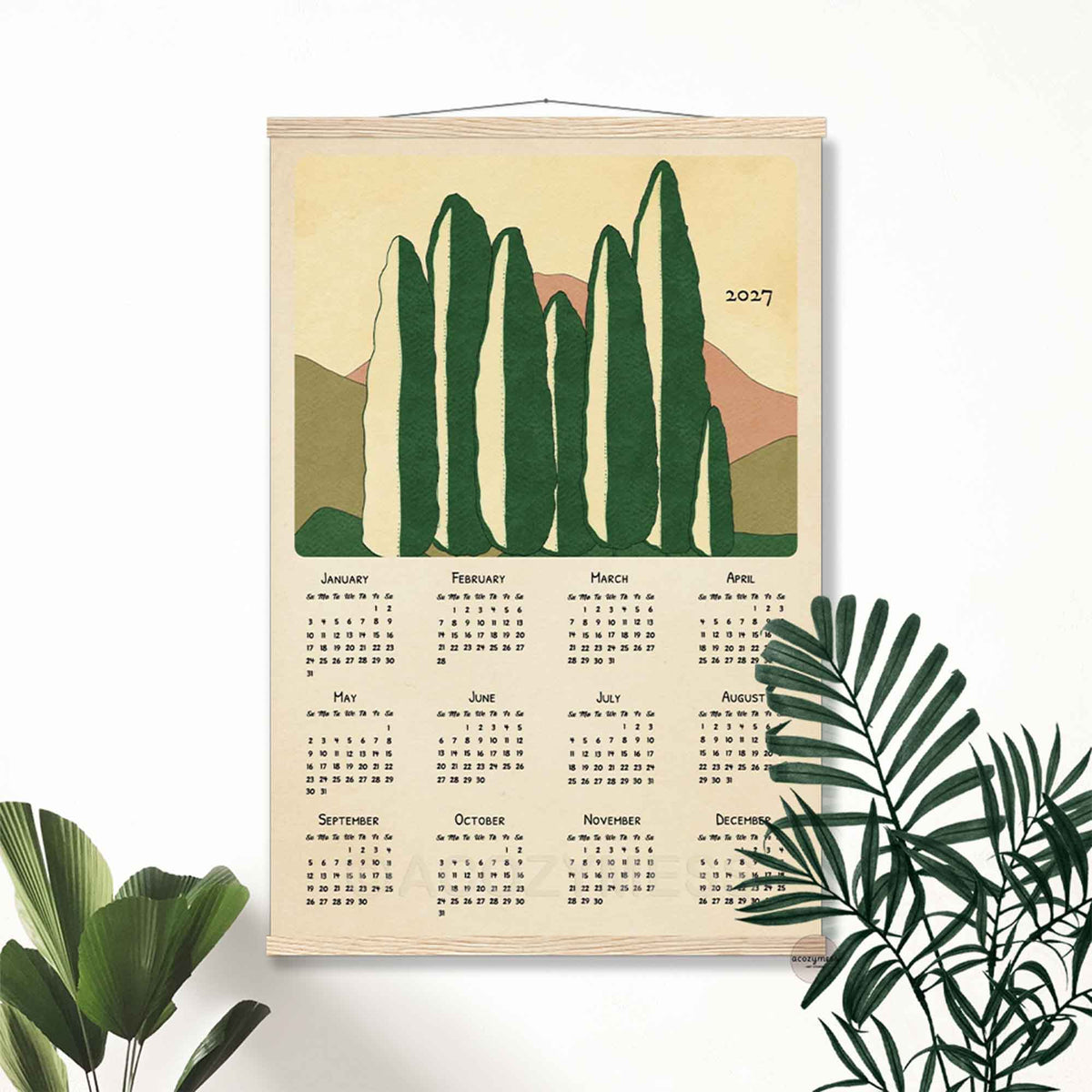 2027 mid-century tree landscape calendar in a light wood hanger frame on a white wall with tropical plants