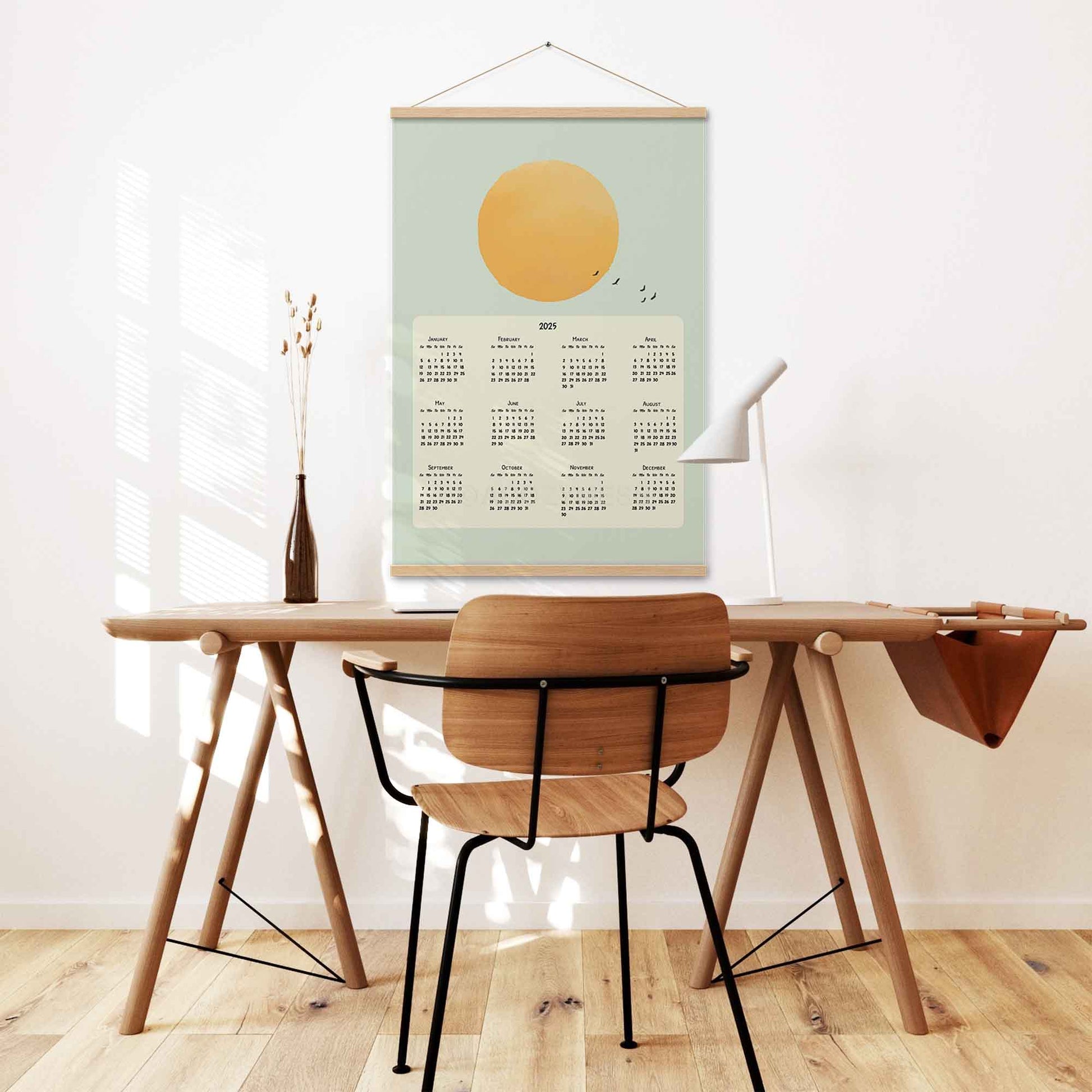 2025 yearly calendar poster featuring a sun illustration in a blue sky with flying birds,  with oak hanger on a desk wall.