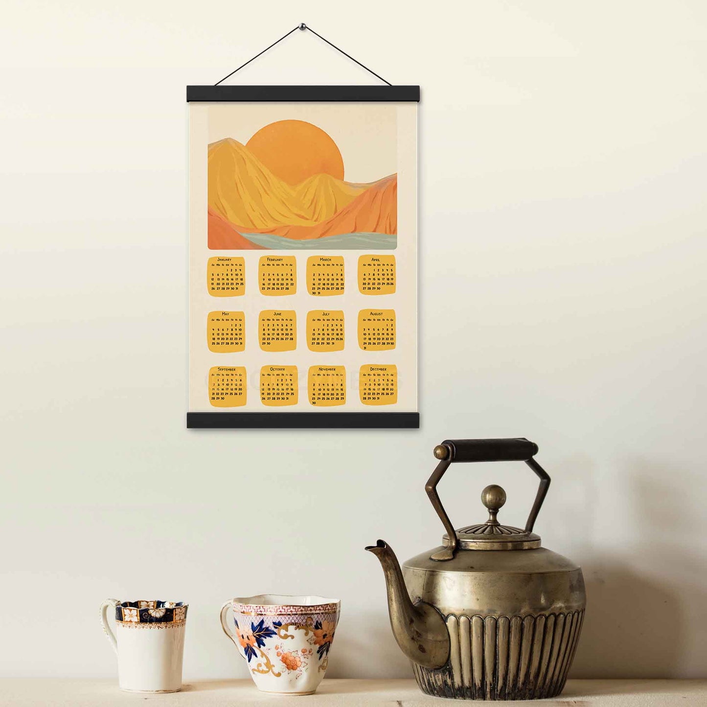 2025 illustrated calendar featuring a serene mountain view in yellow and orange tones, displayed with an black hanger on a kitchen wall, perfect for adding warmth and charm.