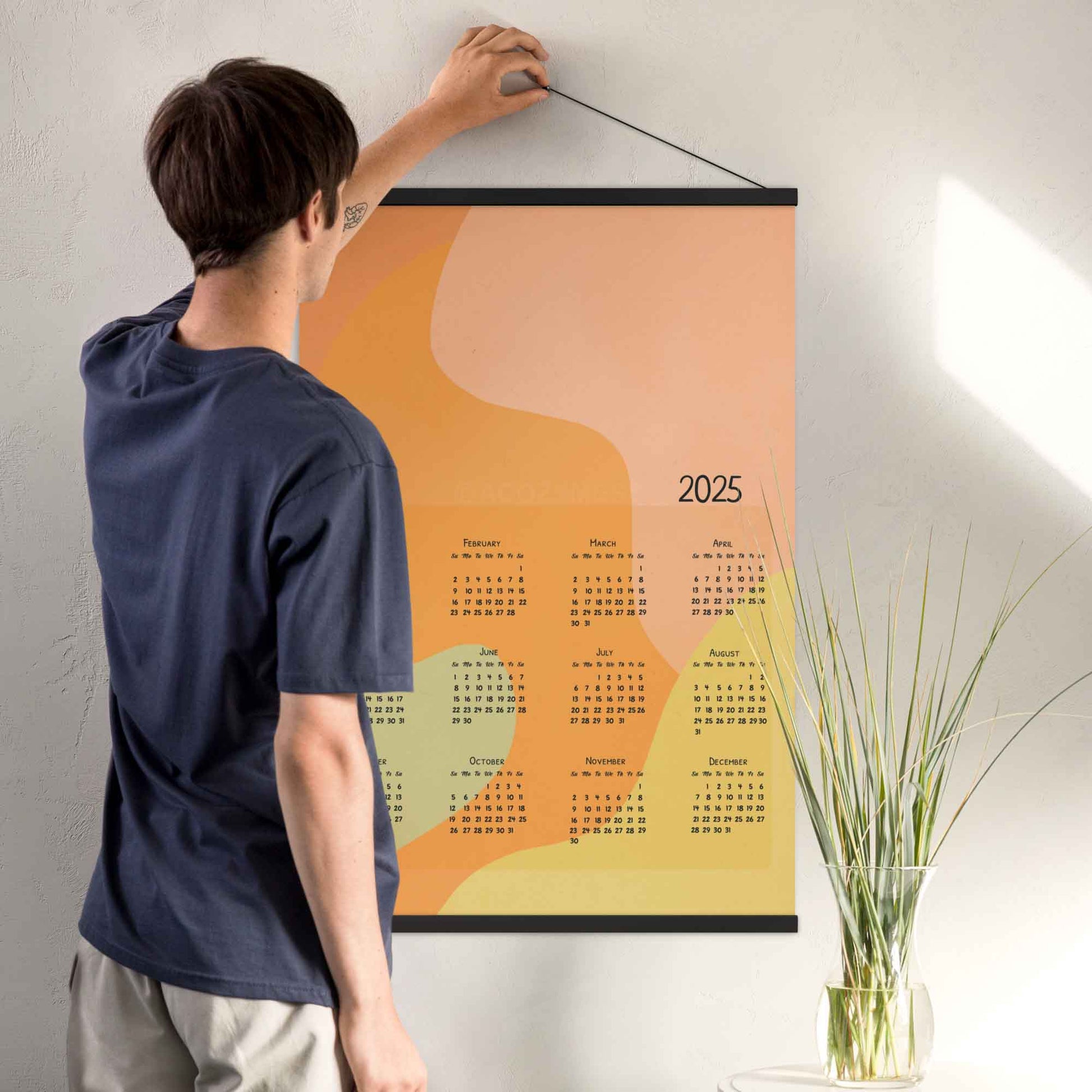 2025 yearly calendar poster with an abstract design in pastel colors, displayed with an black hanger on a living room wall.