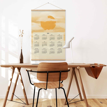 2025 yearly wall calendar featuring a sun and clouds illustration on a muted yellow background, adding warmth to the design, displayed in a oakwood hanger frame on a desk wall.