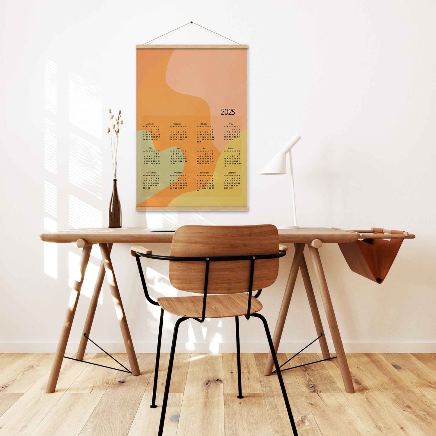 2025 yearly calendar poster with an abstract design in pastel colors, displayed with an wood hanger on a desk wall.