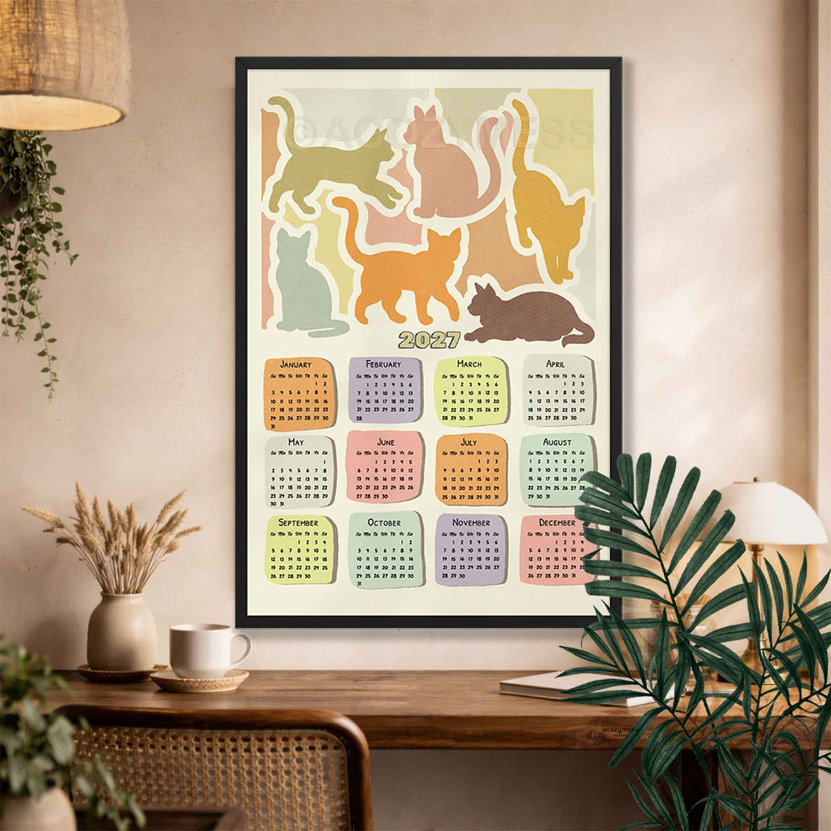 Black framed 2027 cat calendar poster with pastel cat silhouettes above a wooden desk with a coffee cup, dried wheat vase and rattan chair