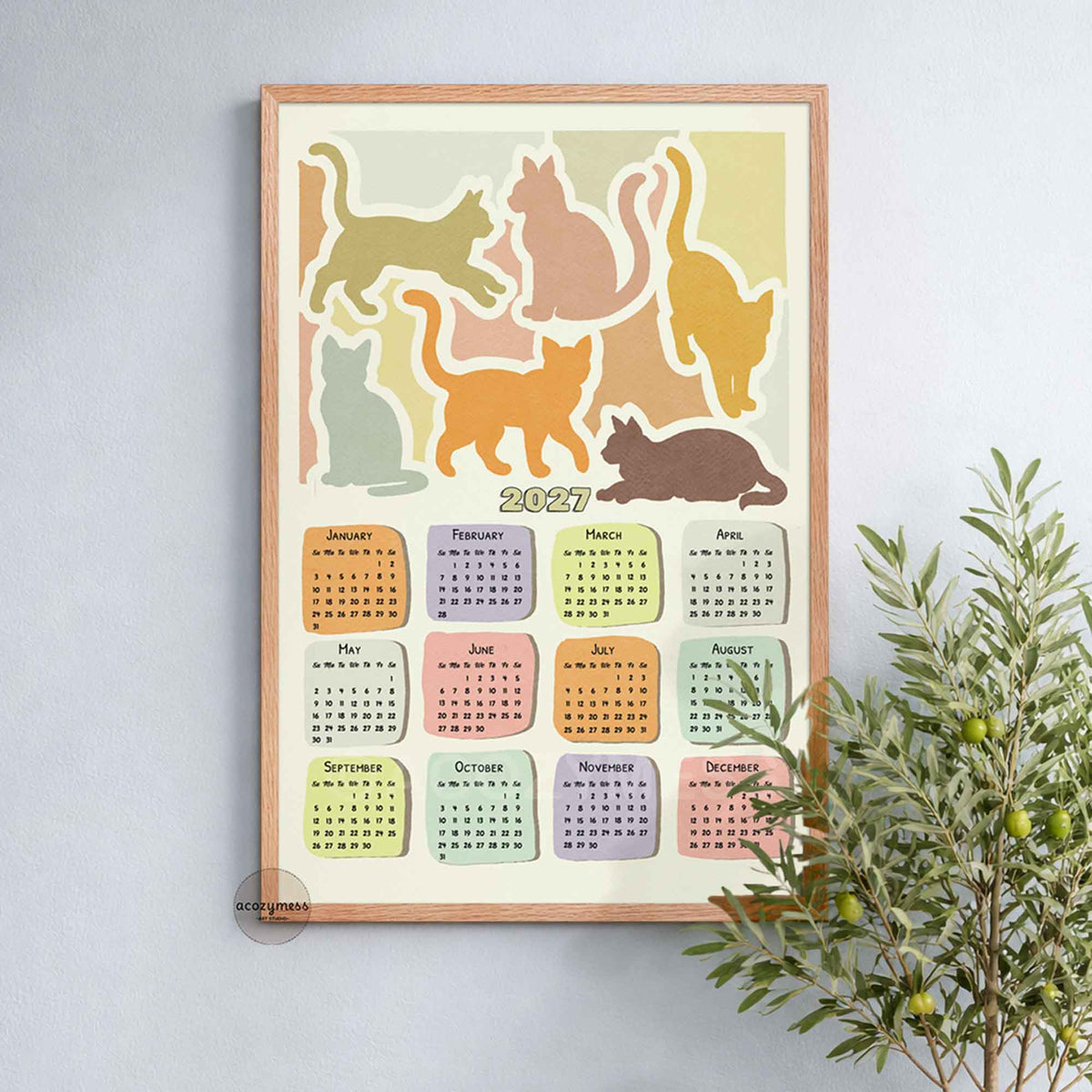 Oak framed 2027 yearly calendar cat art print on a soft blue wall beside a leafy olive plant