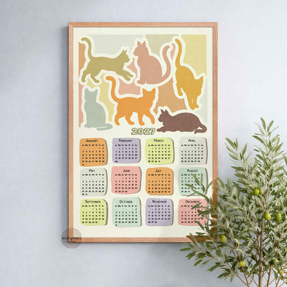 Oak framed 2027 yearly calendar cat art print on a soft blue wall beside a leafy olive plant