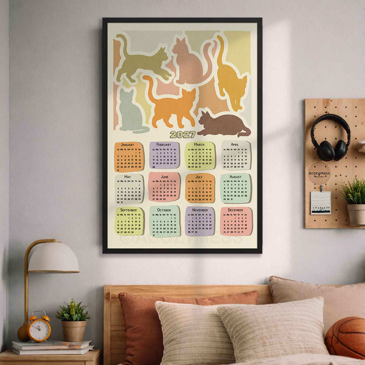 Black framed 2027 cat calendar wall art above a bed with cozy pillows, a lamp and a pegboard with headphones