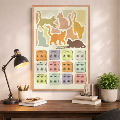 Oak framed 2027 cat silhouette calendar poster above a desk with a black lamp, books and a potted plant