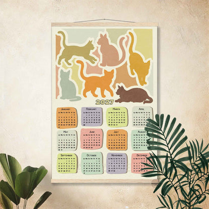 2027 cat calendar poster in a wooden hanger frame on a warm cream wall with palm leaves