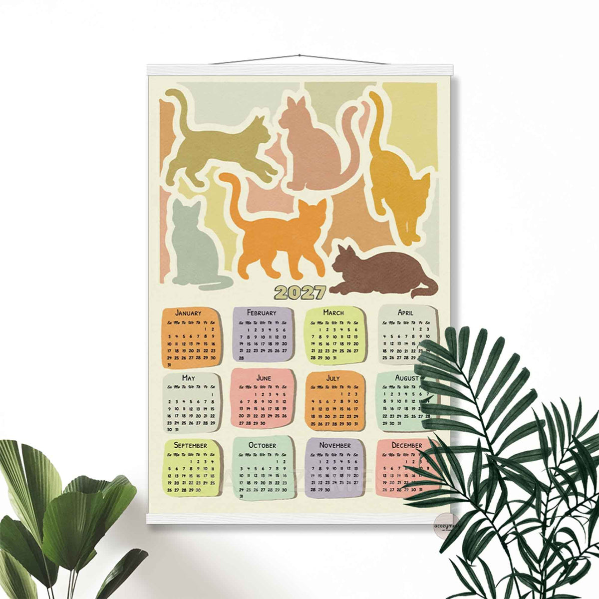 2027 yearly cat calendar print in a light wooden hanger frame on a white wall with tropical plants