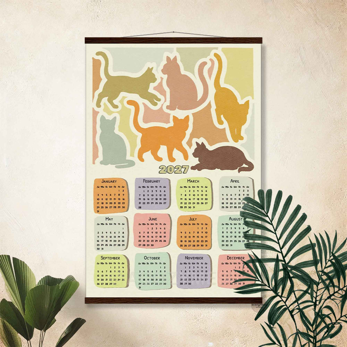 2027 cat calendar poster in a dark wooden hanger frame on a beige wall with palm leaves