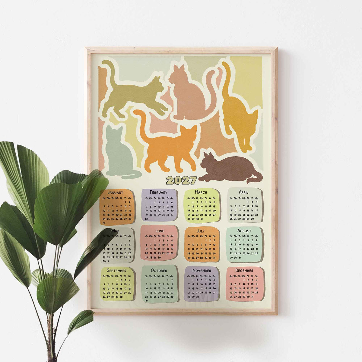 Oak framed 2027 cat calendar wall art on a white wall beside a large green plant