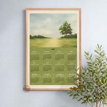 Oak framed 2027 landscape calendar poster with a green meadow and lone tree on a soft blue wall beside an olive plant