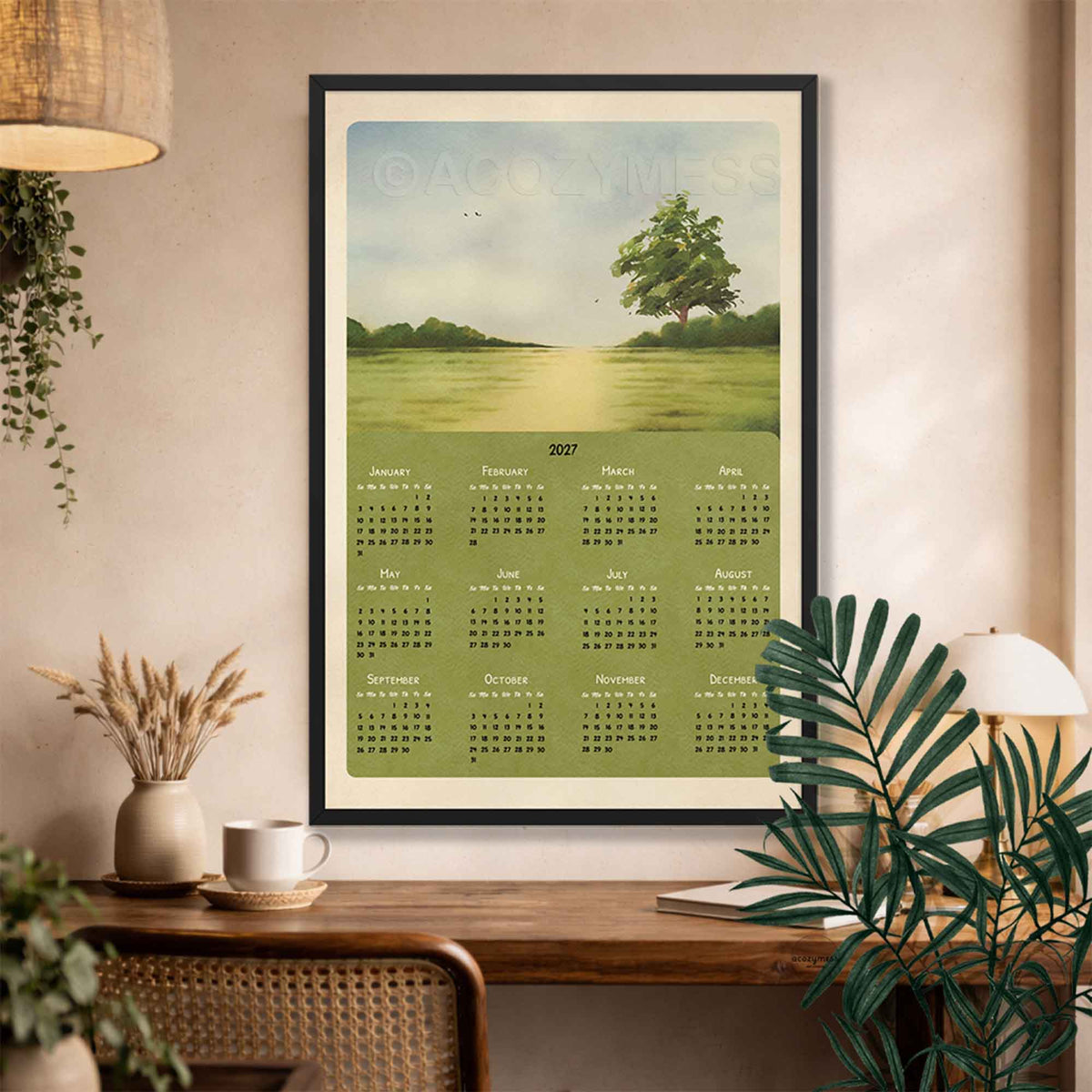 Black framed 2027 landscape calendar above a wooden desk with a coffee mug, dried grasses and a rattan chair