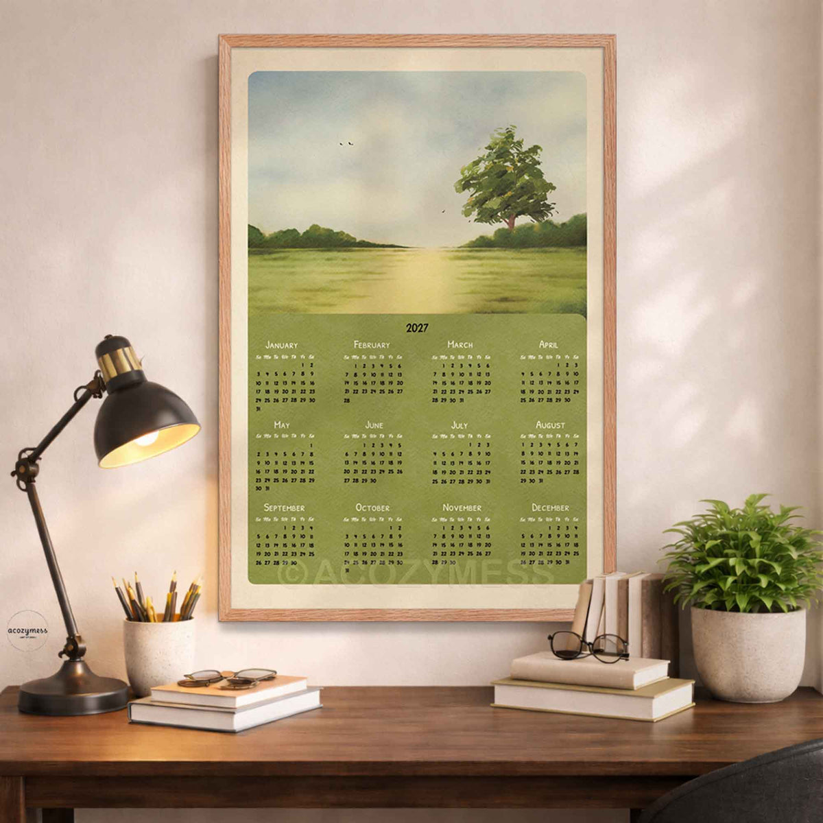 Oak framed 2027 green meadow calendar above a desk with a black lamp, books and a potted plant