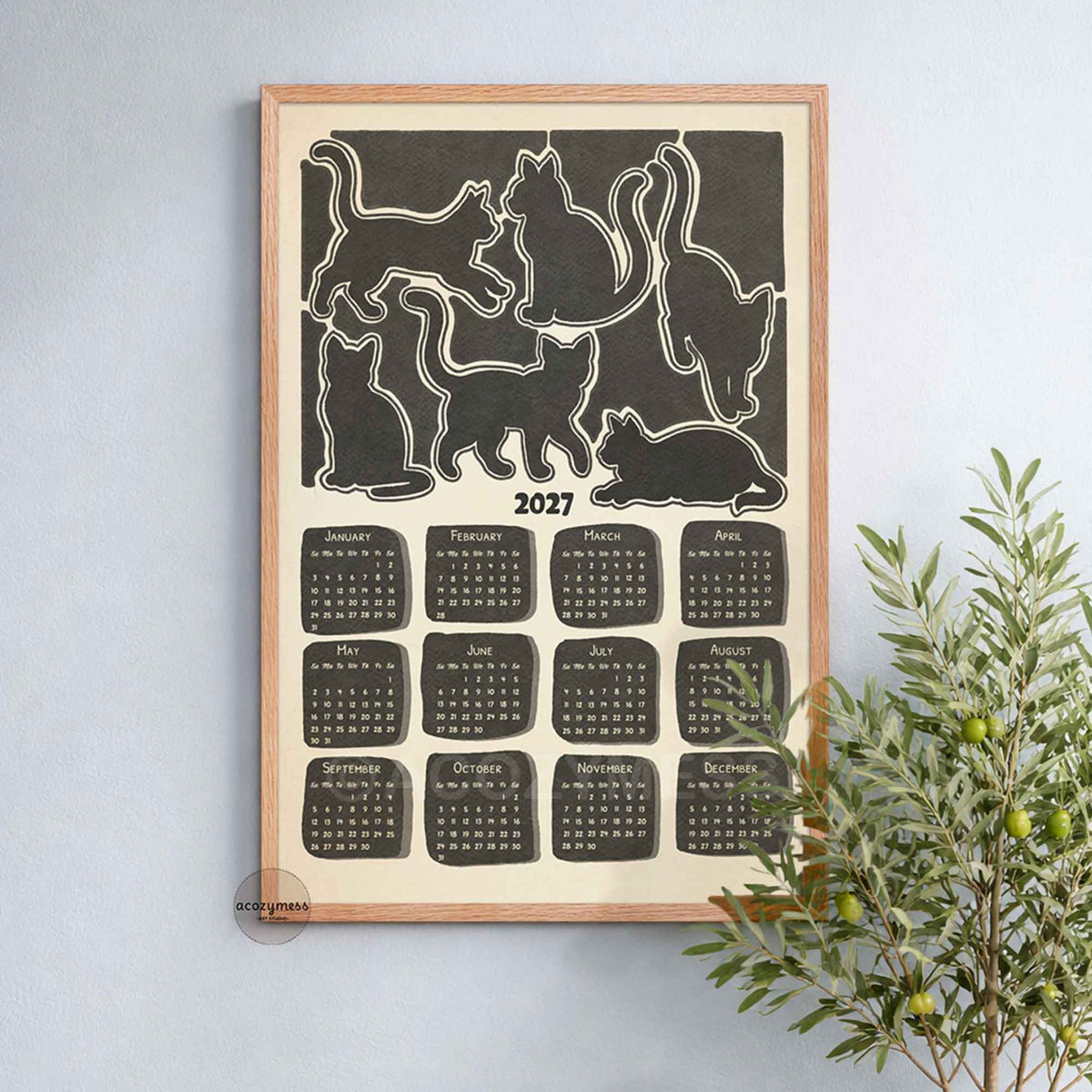 Oak framed 2027 monochrome cat calendar poster on a soft blue wall beside a leafy olive plant