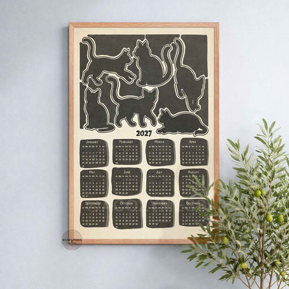 Oak framed 2027 monochrome cat calendar poster on a soft blue wall beside a leafy olive plant