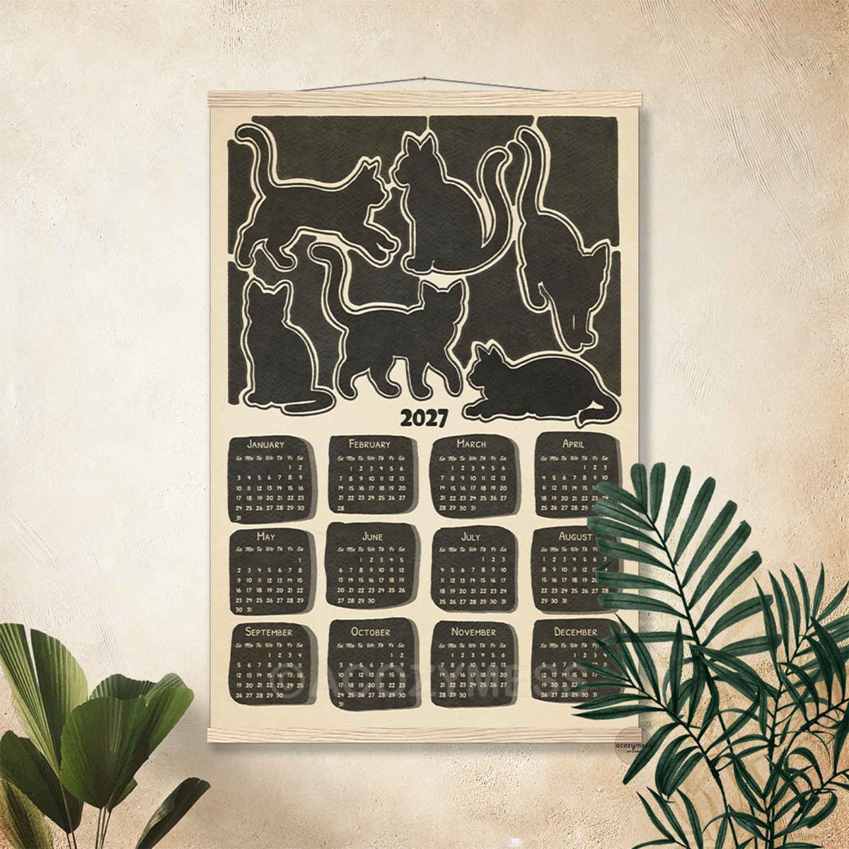 2027 monochrome cat calendar print in a light wooden hanger frame on a cream wall with tropical plants