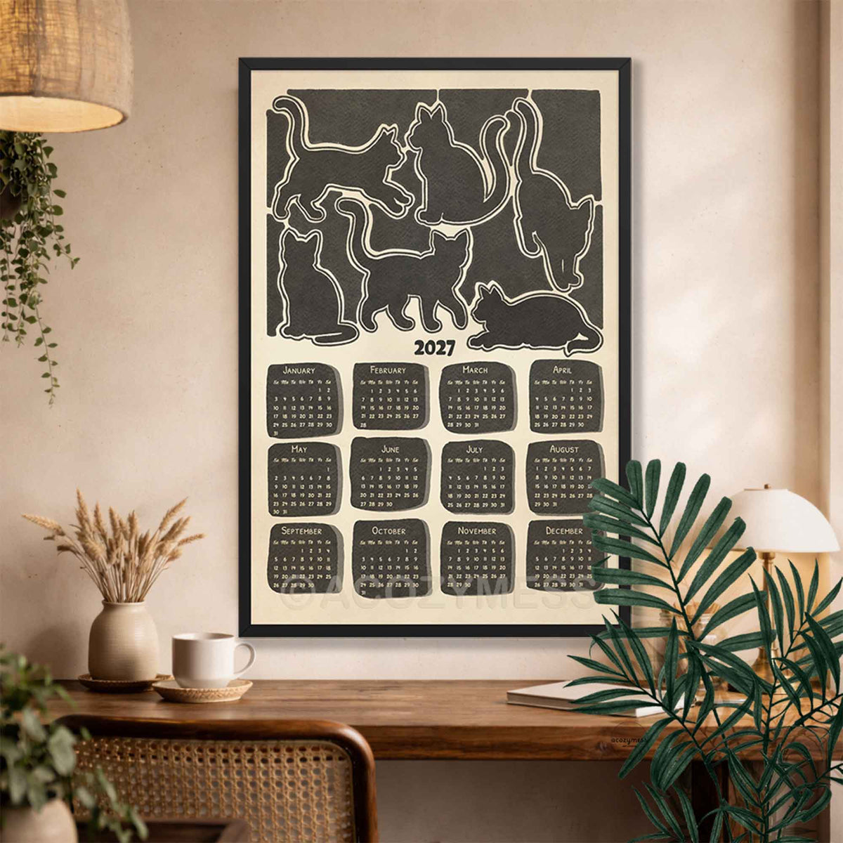 Black framed 2027 cat calendar wall art above a wooden desk with a coffee cup, dried flowers and a rattan chair