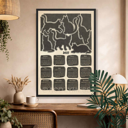 Black framed 2027 cat calendar wall art above a wooden desk with a coffee cup, dried flowers and a rattan chair