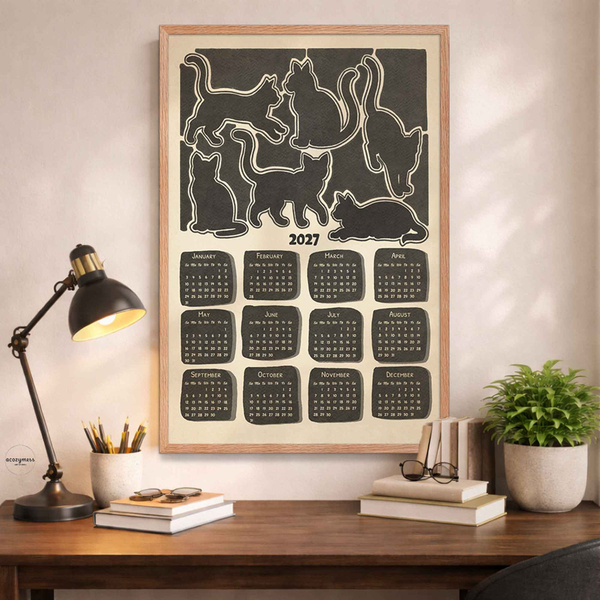 Oak framed 2027 minimalist cat calendar poster above a desk with a black lamp, books and a potted plant