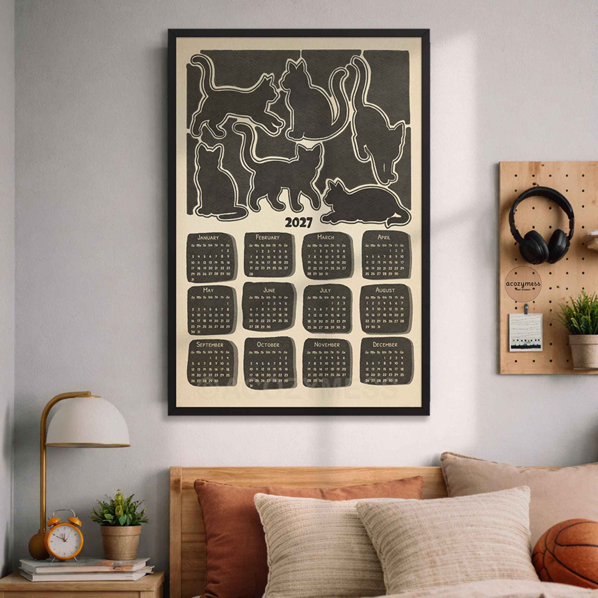 Black framed 2027 cat silhouette calendar above a bed with cozy pillows, a lamp and a pegboard with headphones