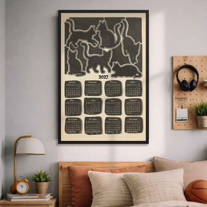 Black framed 2027 cat silhouette calendar above a bed with cozy pillows, a lamp and a pegboard with headphones