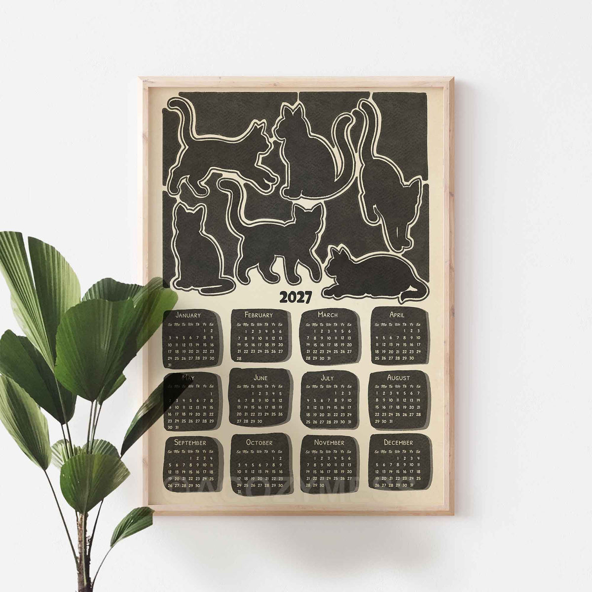 Light wood framed 2027 monochrome cat calendar on a white wall beside a large green plant