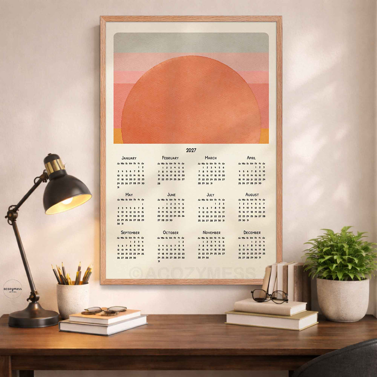 Oak framed 2027 sun calendar poster above a wooden desk with a black lamp, books and a potted plant