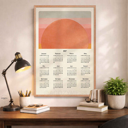 Oak framed 2027 sun calendar poster above a wooden desk with a black lamp, books and a potted plant