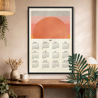 Black framed 2027 boho sunset calendar above a desk with a rattan chair, dried grasses and palm leaves