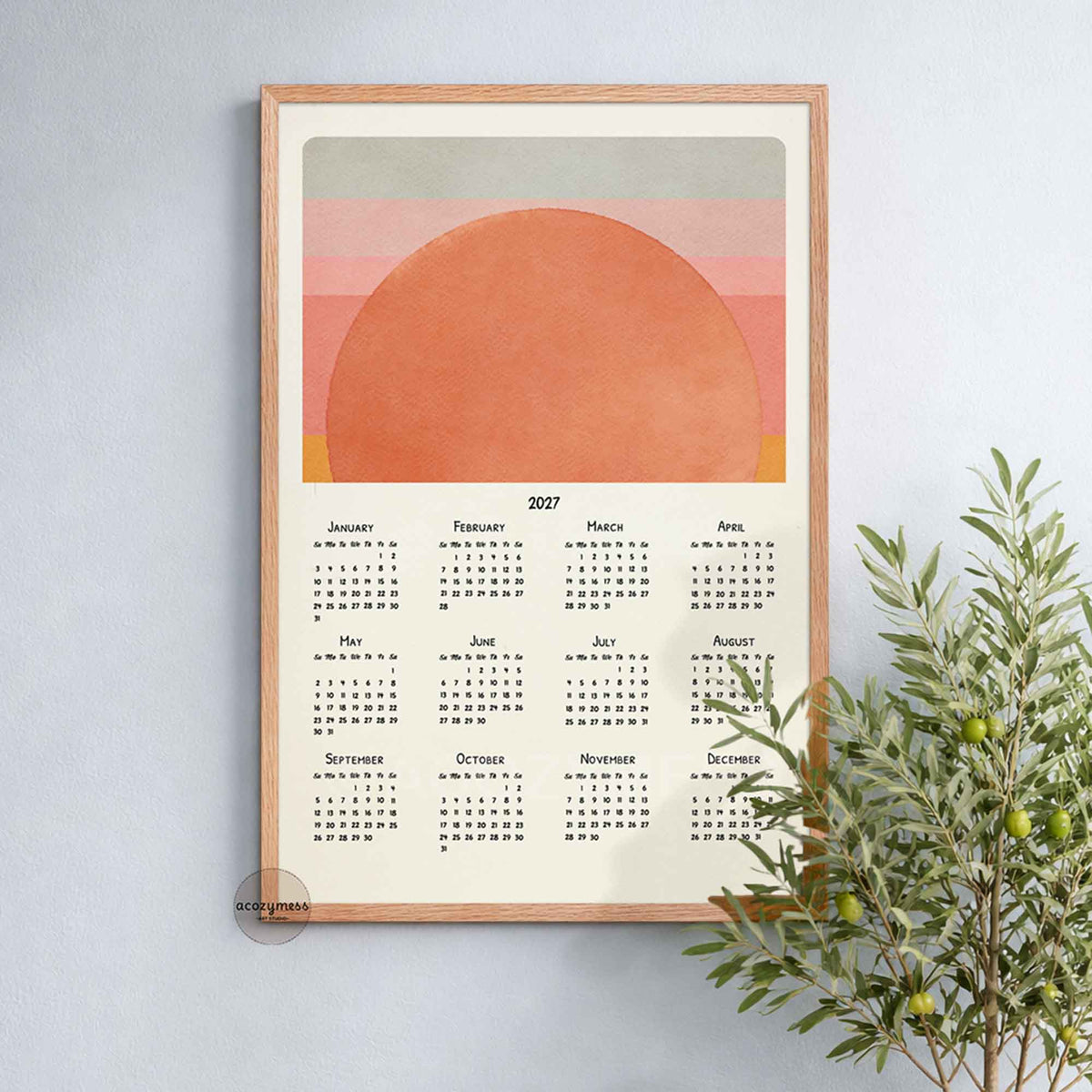 Oak framed 2027 sun calendar print on a soft blue wall beside an olive tree