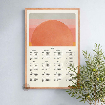 Oak framed 2027 sun calendar print on a soft blue wall beside an olive tree
