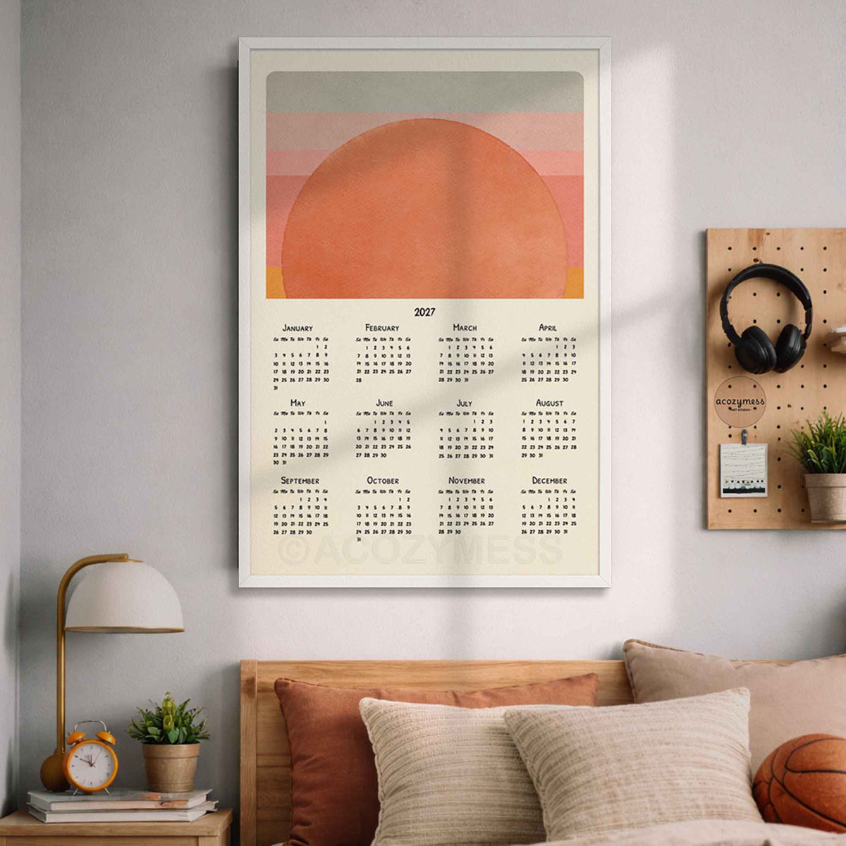 White framed 2027 mid-century sun calendar above a bed with pillows and a wooden pegboard