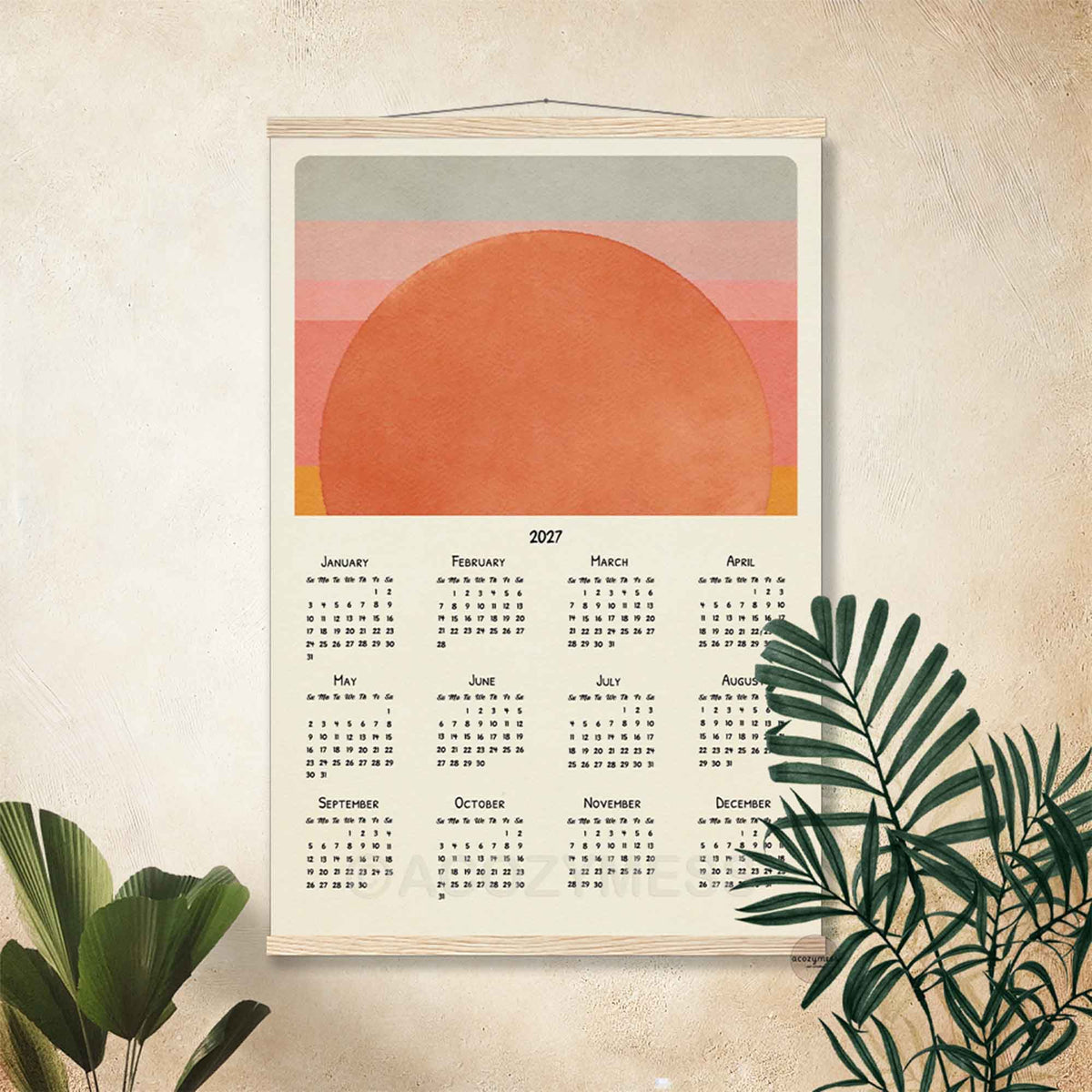 2027 sun calendar print in a light wood hanger frame on a textured beige wall with palm leaves