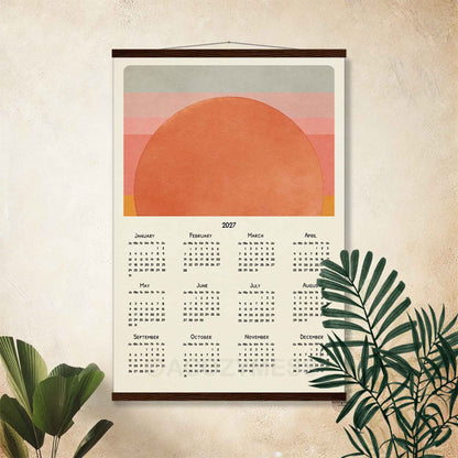 2027 sunset wall calendar in a dark wood hanger frame on a textured beige wall with palm leaves