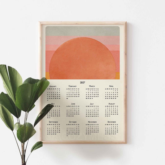 Light wood framed 2027 sun calendar poster on a white wall beside a large green plant