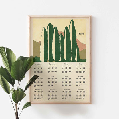 Light oak framed 2027 cypress trees calendar poster on a white wall beside a green fan palm plant