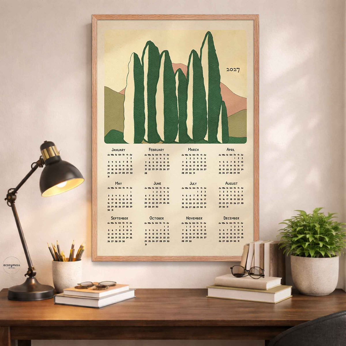 Light oak framed 2027 cypress trees calendar poster above a wooden desk with a black lamp, books and a potted plant