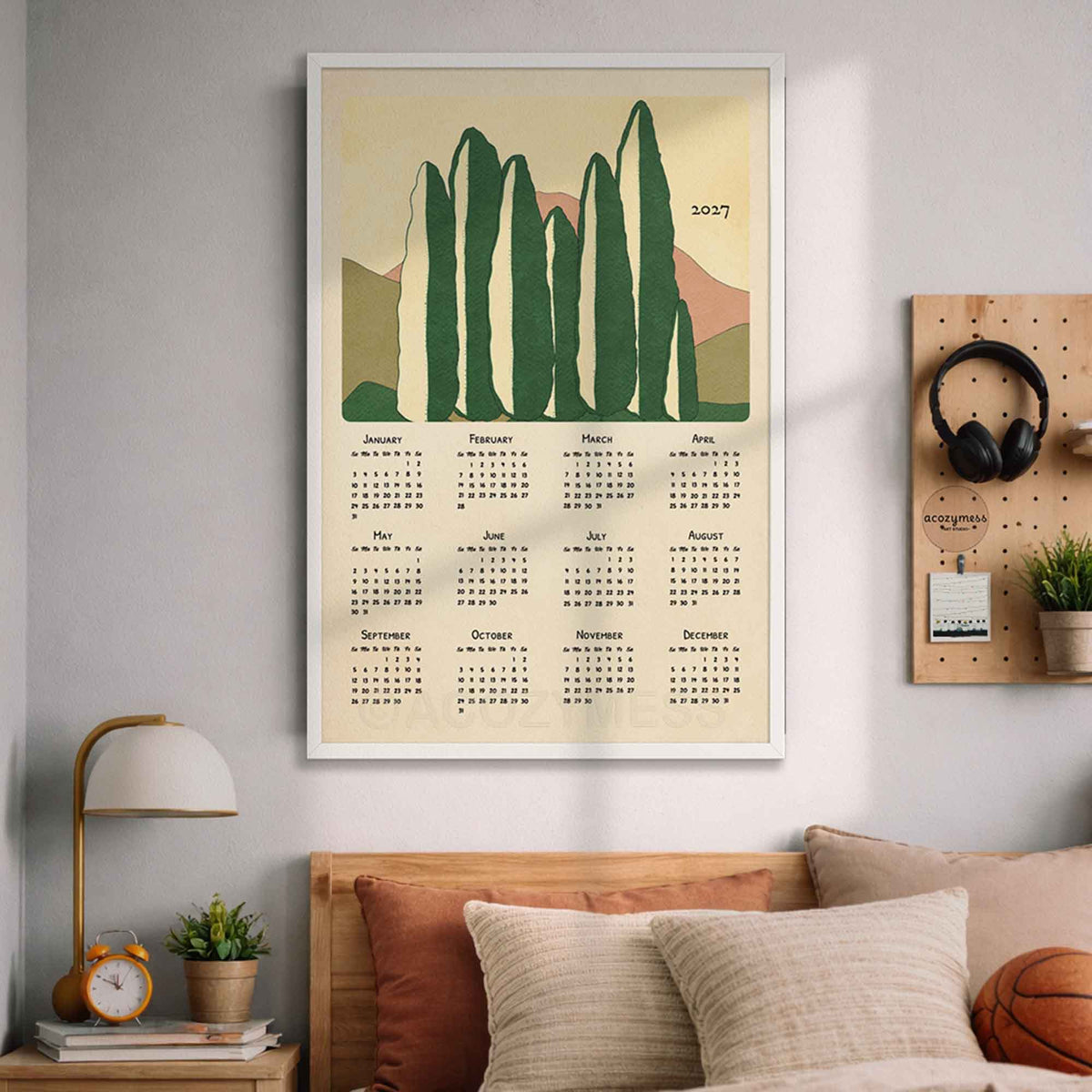 White framed 2027 green cypress tree calendar print on a soft blue wall above a bed with pillows