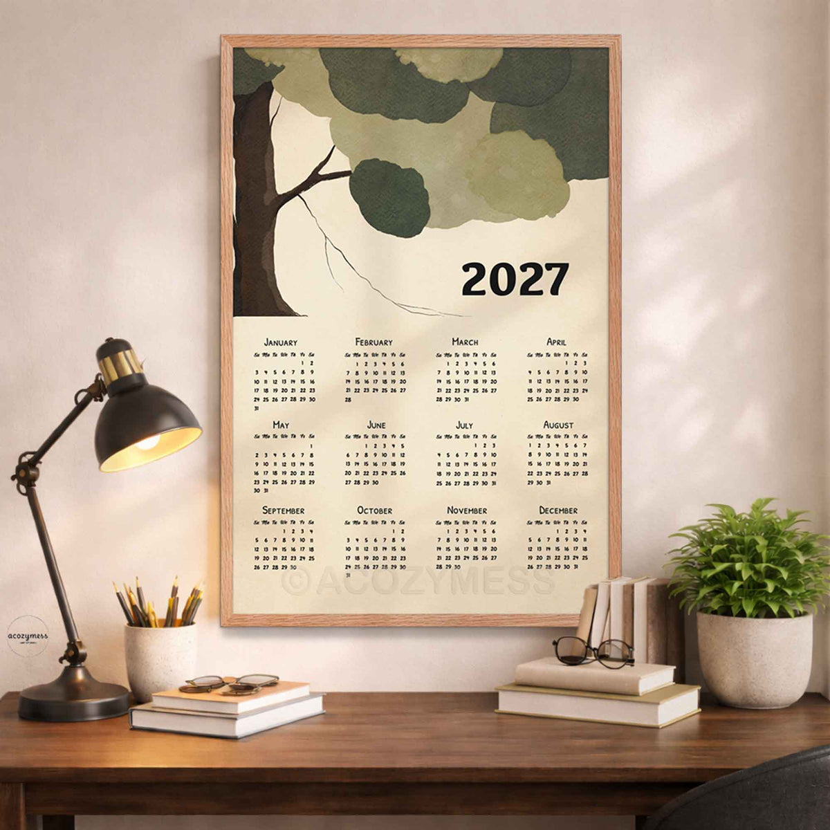 Oak framed 2027 tree calendar print above a wooden desk with a black lamp, books and a potted plant