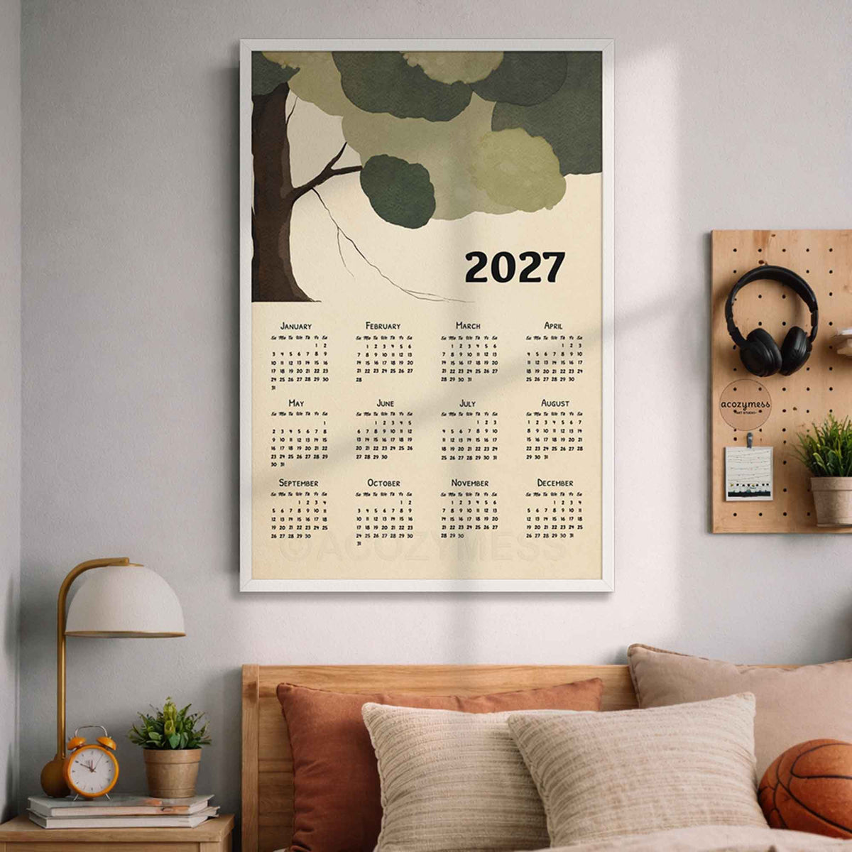 White framed 2027 green tree wall calendar above a bed with pillows and a wooden pegboard