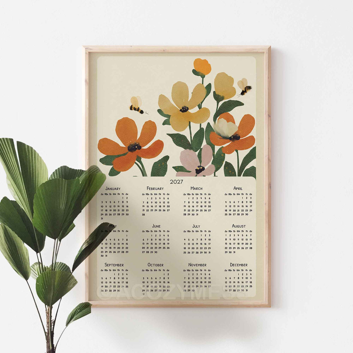 Oak framed 2027 floral calendar poster with orange flowers and bees on a white wall beside a fan palm plant