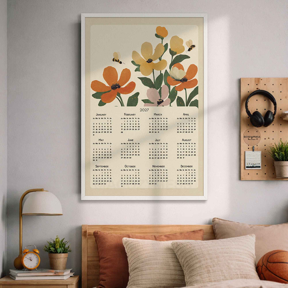 White framed 2027 orange floral wall calendar above a bed with pillows and a wooden pegboard