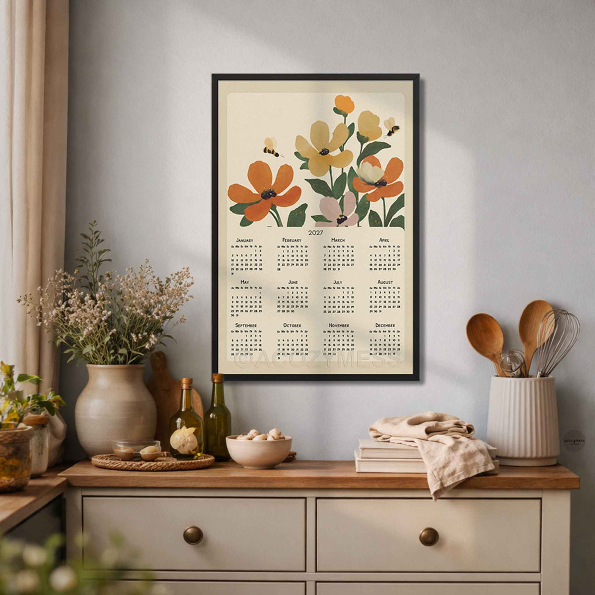Black framed 2027 flowers and bees calendar poster above a cream dresser with vases and kitchen utensils