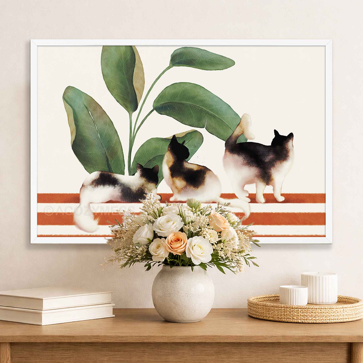 Framed artwork of three cats on a striped bench with plants, displayed above a table with a vase and flowers.