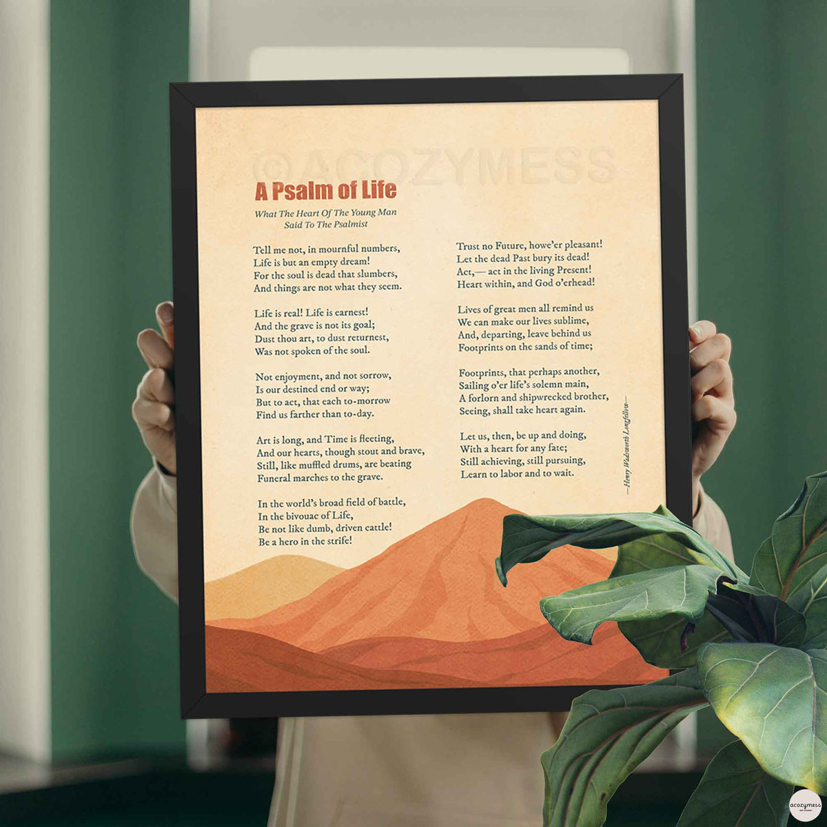 Hands holding a black framed A Psalm of Life poem print with burnt orange mountain art against a green wall