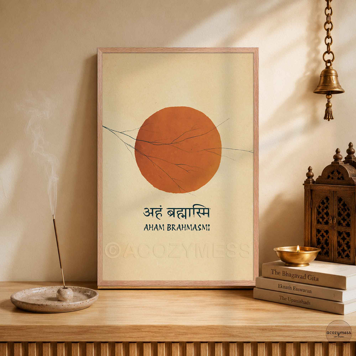 A framed art print featuring a large sun and the Sanskrit/Hindi text 'Aah Brahmasi' with 'Aham Brahmasi!' underneath, displayed on a wooden shelf in a styled room setting with a brass incense holder, books, and a hanging brass bell.