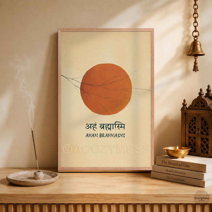 A framed art print featuring a large sun and the Sanskrit/Hindi text 'Aah Brahmasi' with 'Aham Brahmasi!' underneath, displayed on a wooden shelf in a styled room setting with a brass incense holder, books, and a hanging brass bell.