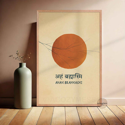 A framed minimalist art print leaning against a wall on a wooden floor. The print features a large orange sun art  with thin dark branches on a warm cream background, with the text ''AHAM BRAHMASI' printed below.